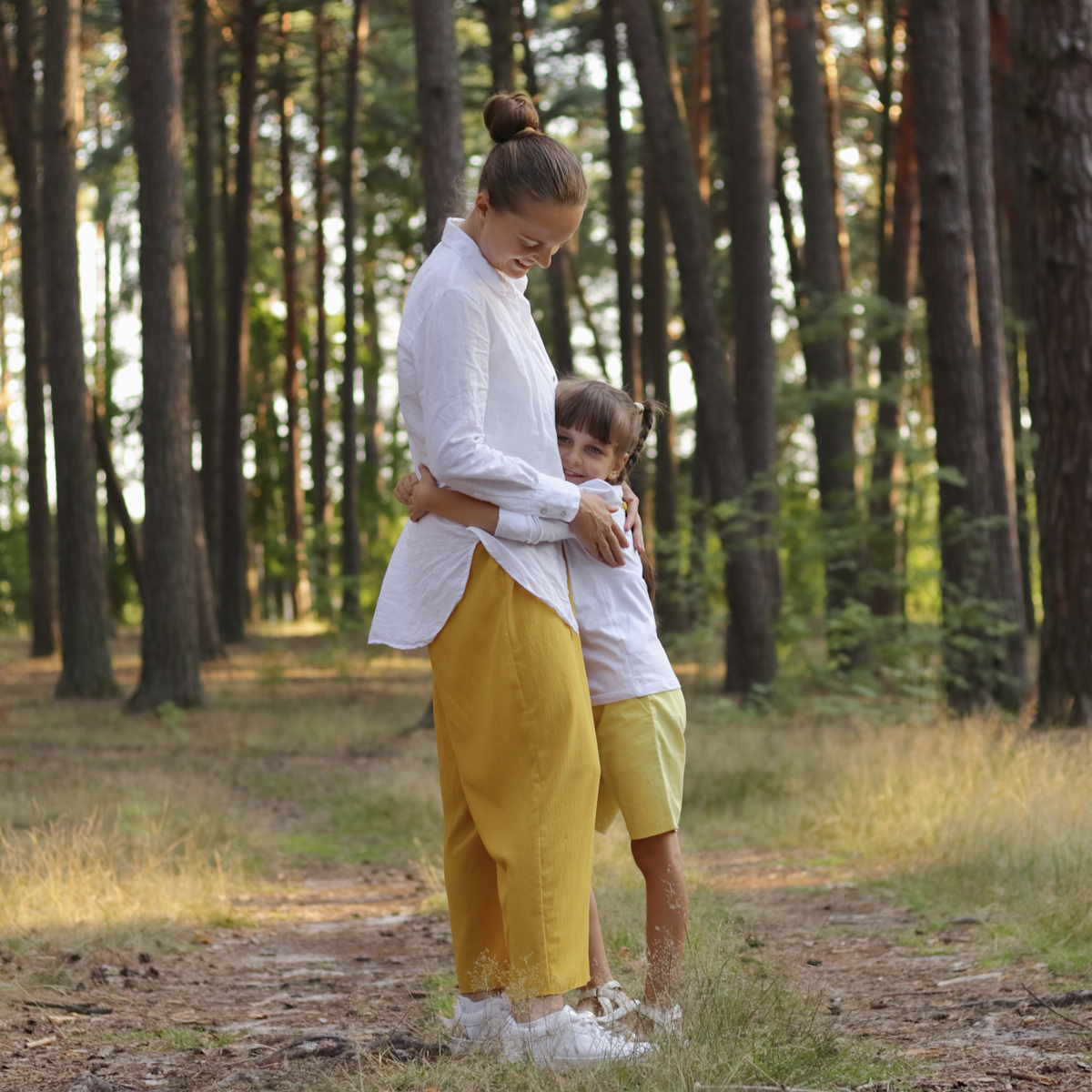 Full length portrait of happy mother standing and hugging her cute charming daughter in the middle of the forest, family walking in wood, breathing fresh air, enjoying nature.